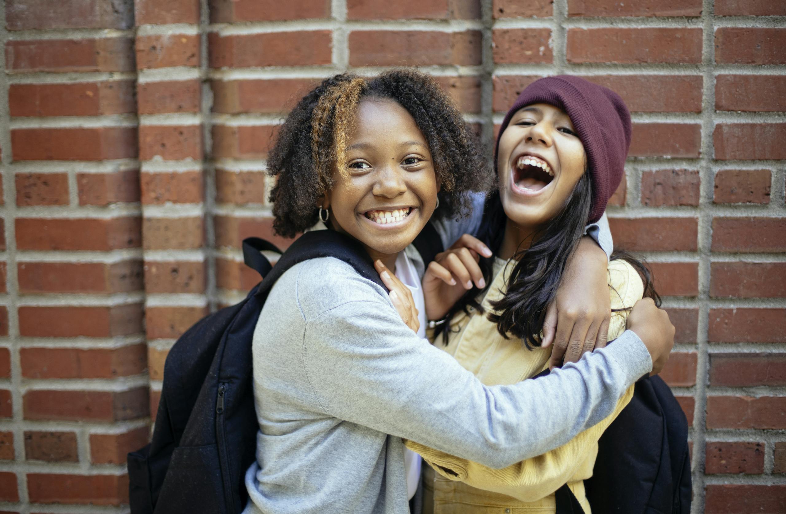 Two joyful girls share a warm embrace against a brick wall, showcasing friendship and happiness.